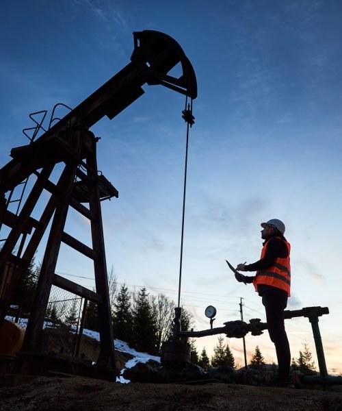 Side view snapshot of an oil engineer wearing orange vest and a helmet, looking at the oil rig and making some notes against beautiful evening sky. Concept of oil extraction and petroleum industry.